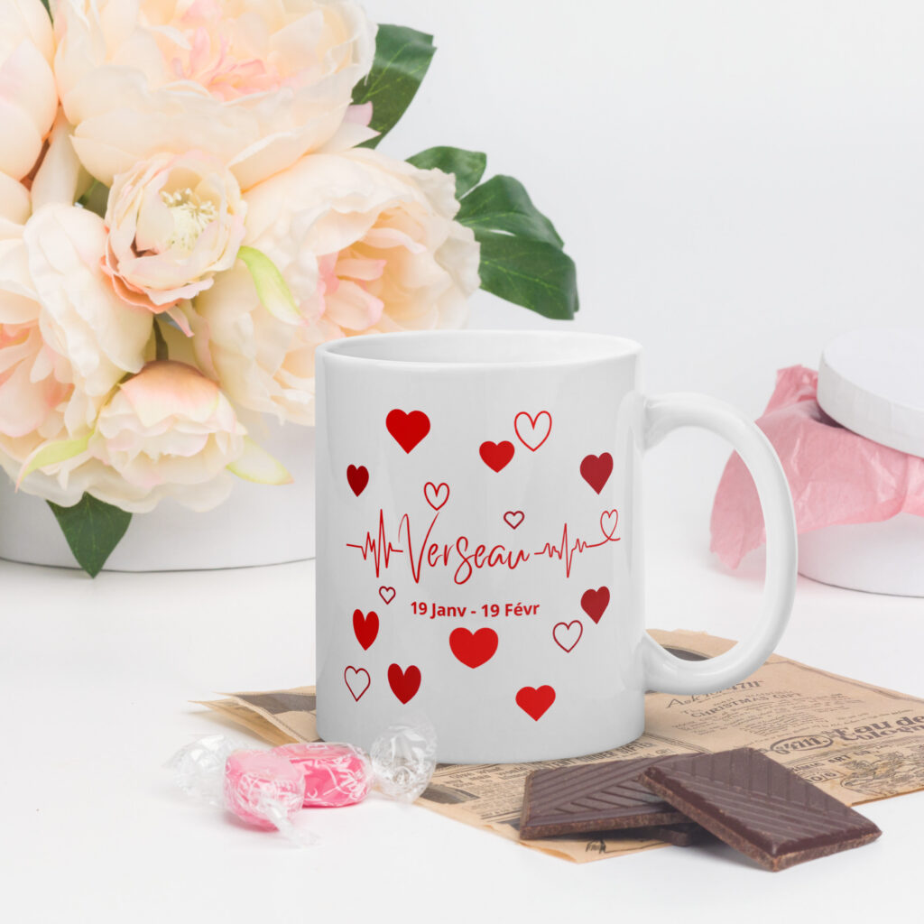 Cadeau original avec Whizaloo : l'Anti-Panne d'idées pour un anniversaire 100% insolite Mug blanc en céramique avec un design astrologique verseau en rouge, entouré de petits cœurs et d'une ligne de battement de cœur. La tasse est posée sur un bureau avec des fleurs roses, du chocolat et des bonbons en arrière-plan.