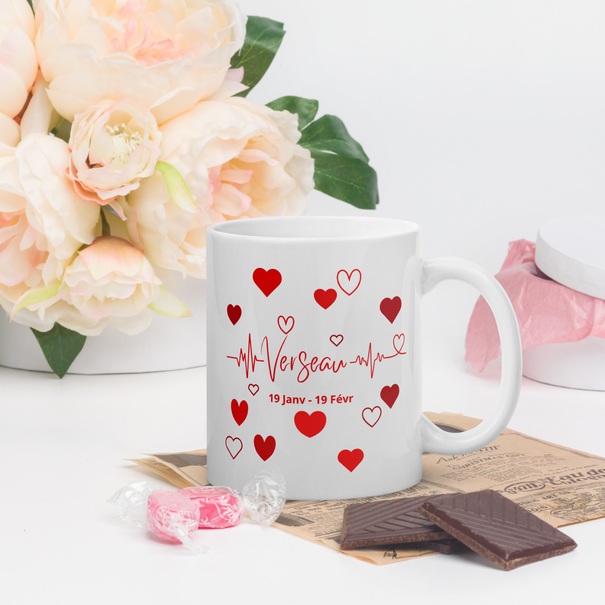 Mug Cœurs ❤️ | Idée Cadeau Romantique pour sa Chérie du signe astrologique Verseau Mug blanc en céramique avec un design astrologique verseau en rouge, entouré de petits cœurs et d'une ligne de battement de cœur. La tasse est posée sur un bureau avec des fleurs roses, du chocolat et des bonbons en arrière-plan.