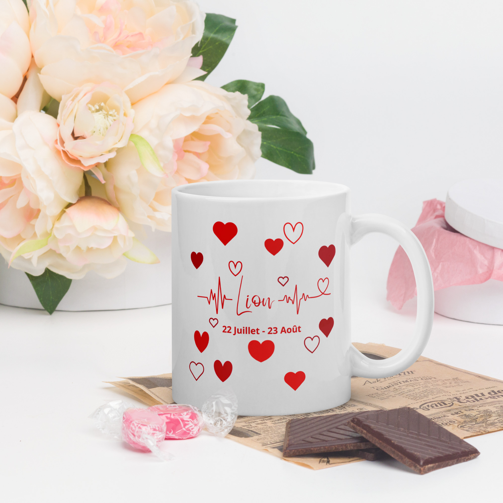 Mug Cœurs ❤️ | Idée Cadeau Romantique pour sa Chérie du signe astrologique Lion Mug blanc en céramique avec un design astrologique lion en rouge, entouré de petits cœurs et d'une ligne de battement de cœur. La tasse est posée sur un bureau avec des fleurs roses, du chocolat et des bonbons en arrière-plan.