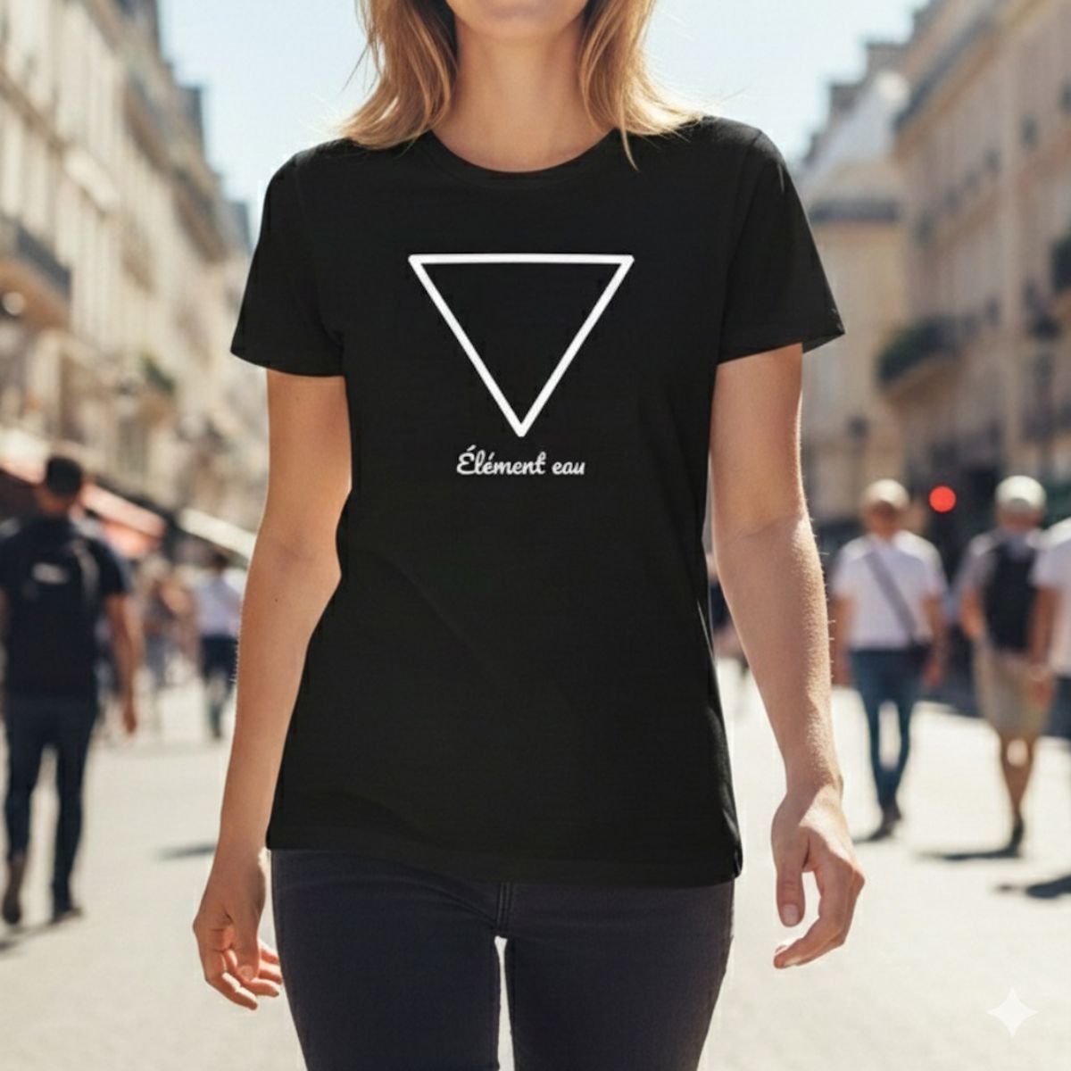 T-Shirt Femme Symbole Élémentaire EAU | symbole ésotérique Portrait d'une jeune femme élégante portant un t-shirt noir tendance à la coupe ajustée, arborant un graphisme blanc du symbole élémentaire de l'eau. Le design ésotérique et minimaliste est complété par l'inscription calligraphiée "Élément eau". Elle marche dans une rue urbaine lumineuse, illustrant un style fashion décontracté et moderne.