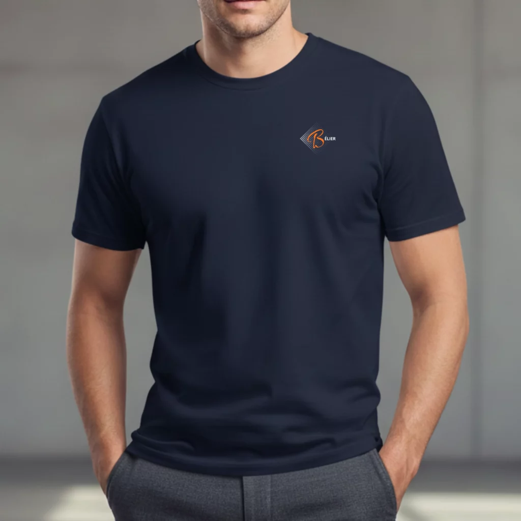Pourquoi Whizaloo a choisi le minimalisme plutôt que le folklore Un homme portant un t-shirt bleu marine à manches courtes et col rond. Sur le côté gauche de la poitrine se trouve un logo discret composé d'un losange graphique gris, d'une lettre "B" orange stylisée et du mot "BELIER" en blanc. L'homme porte un pantalon gris et a les mains dans les poches.