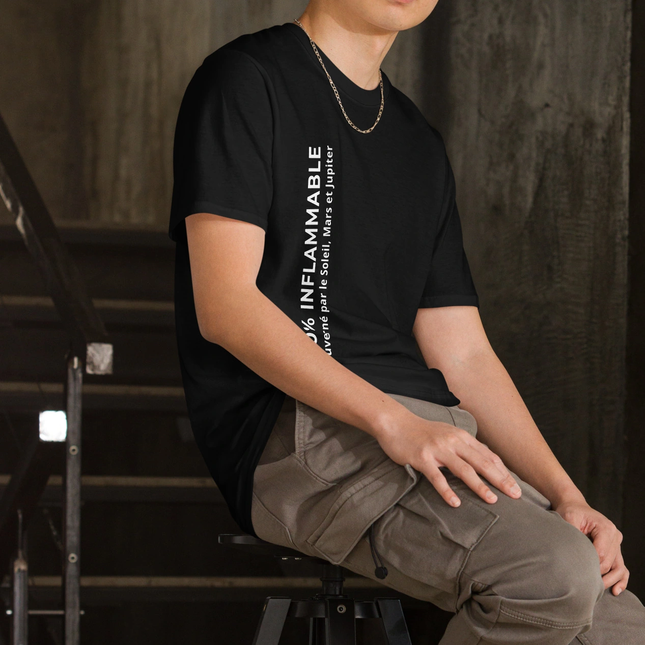 T-shirt Homme Triplicité de Feu « 100% Inflammable – Gouverné par le Soleil, Mars et Jupiter » Photographie d'un homme de trois-quarts, assis sur un tabouret dans un décor industriel aux tons sombres. Il porte un t-shirt noir à manches courtes et un pantalon cargo beige. Sur le côté du t-shirt, on aperçoit une inscription verticale blanche : en grandes lettres capitales « 100% Inflammable », accompagnée de la phrase plus petite « Gouverné par le Soleil, Mars et Jupiter ». L'homme porte également une fine chaîne dorée autour du cou.