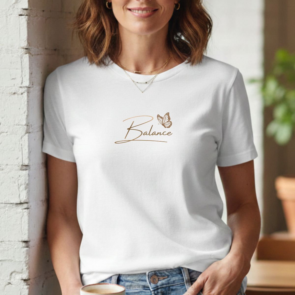 T-shirt Astro Graphique & Papillon | Design XXL - Femme Signe Balance Portrait lifestyle d'une femme souriante portant un t-shirt blanc en coton au design du signe astral balance. Le motif, imprimé en grand format sur la poitrine, présente le nom du signe en typographie fine dorée surmonté d'un papillon délicat. Elle est appuyée contre un mur en briques blanches dans un intérieur lumineux et épuré, agrémenté d'une plante verte en arrière-plan. L'ensemble dégage une esthétique Clean Girl et Basique Chic.