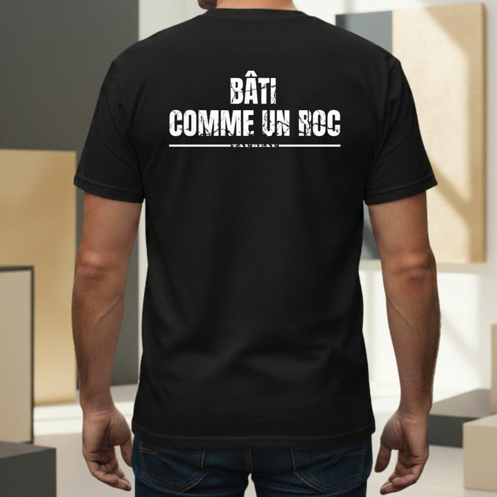 T-shirt noir pour homme avec inscription blanche 'BÂTI COMME UN ROC' et mention 'taureau' soulignée. Design graphique au style usé/vintage centré dans le dos, porté par un modèle masculin dans un cadre extérieur moderne.