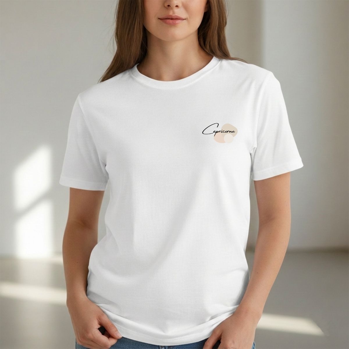 Une femme portant un T-shirt blanc minimaliste avec le mot « Capricorne » inscrit en cursive noire sur la poitrine, accompagné de formes abstraites beiges et neutres.